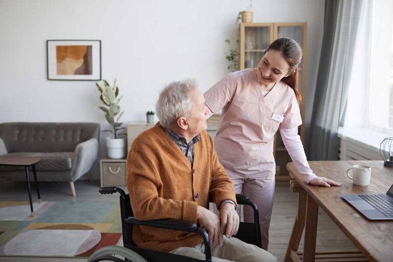 Caregiver helps an older resident stand safely while another staff member checks a care chart nearby.